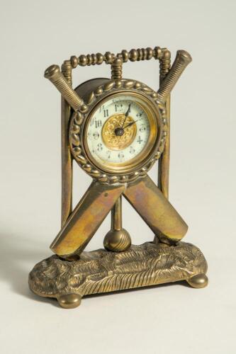 Cricket clock. A Victorian brass mantel clock. The case in the form of a set of stumps with crossed bats and ball on an oval naturalistic case base with bun feet. Dial with gilded centre. Approximately 6.5&rdquo; high by 5&rdquo; wide. British Union Clock Company sta