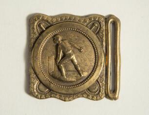 Cricket belt buckle. An almost square shaped brass belt buckle with central figure of a bowler at the wicket to central circle with crossed bat decoration to borders. Circa 1870. Approximately 2.25&rdquo; square. Good condition