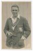 Maurice William Tate. Sussex & England 1912-1937. Early original sepia real photograph postcard of Tate standing three quarter length wearing cricket attire and M.C.C. tour blazer, pipe in hand. Signed in black ink &lsquo;Maurice Tate&rsquo; to the image. Printed tit