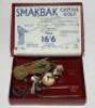 'Smakbak Captive Golf' 1920s. Original golf practice aid comprising golf ball attached to cord with rubber section and two metal staples, lacking tees. In original box with printed illustrated instructions to top and inside of the hinged lid. Splitting to - 2