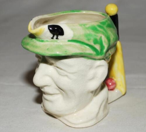 Don Bradman. Toby jug depicting the head of Bradman wearing the green cricket cap as the main vessel and a cricket bat and ball as the handle. White slip and glaze with coloured highlights on the cap and handle. Made by Marutomoware, Japan c1935. Approxim