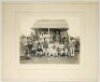 Cricket in the South Africa Second Boer War c.1900. Original sepia photograph of British soldiers and prisoners seated and standing in rows in front of a corrugated pavilion. To verso a handwritten annotation in pencil, and modern printed label laid down,