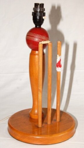 Cricket table lamp. Table lamp by Gray Nicholls comprising a slender wooden baluster pillar surmounted with plastic cricket ball and light fitting, behind a set of cricket stumps and bails. All set on a circular wooden base. One bail detached but both pre