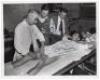 M.C.C. 1950’s. Excellent mono press photograph depicting England masseur Harold Dalton massaging Reg Simpson’s thigh with Simpson laid out on a table shared with crockery and cakes! Len Hutton and Arthur McIntyre are looking on, Hutton with a sandwich in 