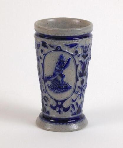 Westerwald stoneware cricket beaker, moulded in relief with three cameo panels of a batsman, after W.G. Grace, in different poses. The body decorated with vine and sprig decoration in cobalt blue on a grey background. Blue rings to rim and to foot. Approx