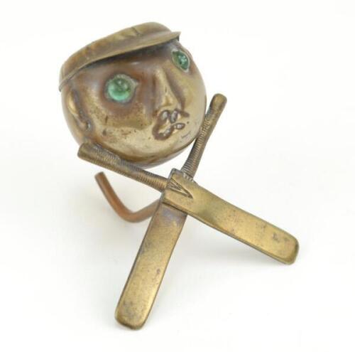 Cricket pin cushion. Edwardian brass metal pin cushion, the cushion on the head of a cricketer wearing cap, the eyes of the cricketer made from coloured green stones. Supported on crossed bat feet. Very unusual. 3” tall. Minor wear otherwise in good condi