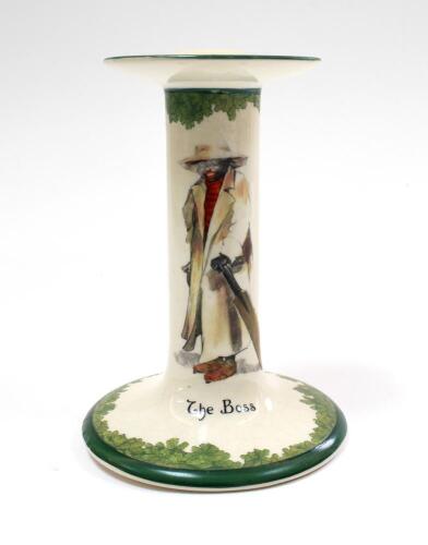 &lsquo;The Boss&rsquo; A very rare Royal Doulton &lsquo;All Black Team&rsquo; series candlestick, entitled &lsquo;The Boss&rsquo;, printed and painted with a boy cricketer in red shirt, wearing an unpire&rsquo;s coat and a floppy hat holding a cricket bat to side Green floral decoration above and