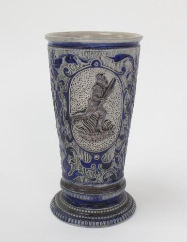 Westerwald stoneware cricket beaker, moulded in relief with three cameo panels of a batsman, after W.G. Grace, in different poses. The body decorated with vine and sprig decoration in cobalt blue on a grey background. Blue rings to rim and to foot. Approx