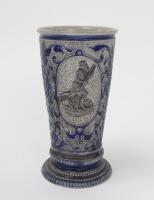 Westerwald stoneware cricket beaker, moulded in relief with three cameo panels of a batsman, after W.G. Grace, in different poses. The body decorated with vine and sprig decoration in cobalt blue on a grey background. Blue rings to rim and to foot. Approx