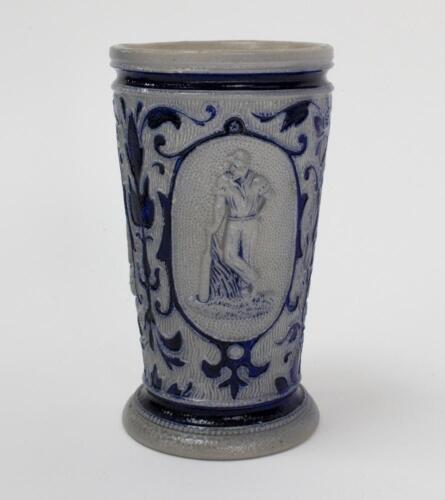 Westerwald stoneware cricket beaker, moulded in relief with three cameo panels of a batsman, after W.G. Grace, in different poses. The body decorated with vine and sprig decoration in cobalt blue on a grey background. Blue rings to rim and to foot. Approx