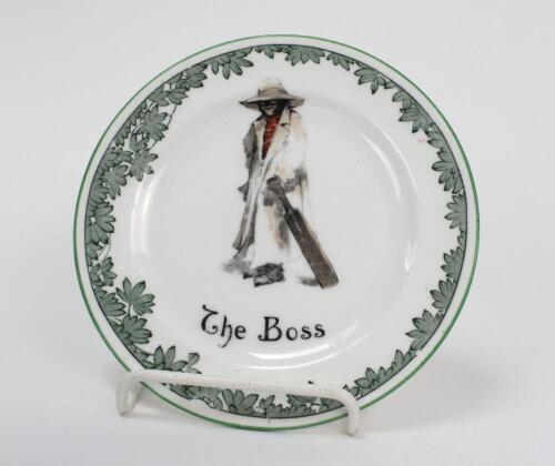 ‘The Boss’ Royal Doulton Black Boy small plate, entitled ‘The Boss’ printed with a boy in red shirt and a floppy hat, wearing an umpires’s coat and holding a bat. Green floral decoration to edge. 5.25” diameter. Doulton backstamp and number ‘E4336’ to bas