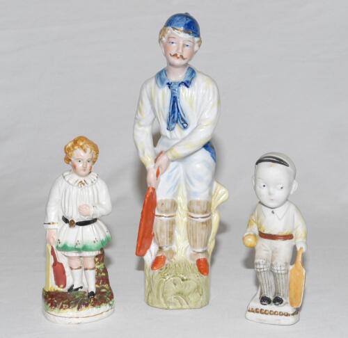 Cricket figures. Selection of three figures. A Continental bisque figure of a batsman, wearing blue cap, yellow shirt, blue cravatte and sash holding a bat to side on a naturalistic oval base. 10” tall. The second a Staffordshire 19th century pottery fig