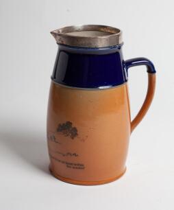 Royal Doulton golf jug in brown with cobalt blue collar and silver rim, decorated with a golfing (cavalier) scene, with motto, ‘A ball is lost if it be not found within five minutes’. 8.75” tall. Doulton mark to base, numbers 8820.BB5. Reg no 525512. Silv