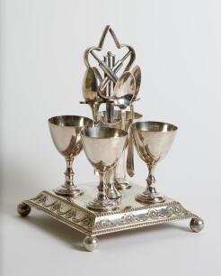Cricket themed egg cup presentation piece. A unique set of four silver-plated egg cups with accompanying spoons on an ornate square based stand with ball feet. The centre piece holding the spoons with crossed bats, ball and wicket decoration. The shield 