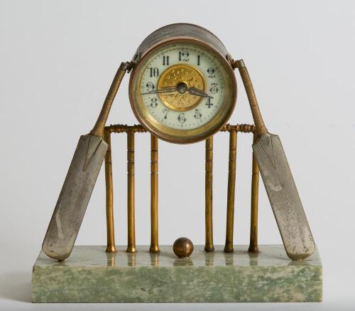 Cricket clock. A Victorian brass mantel clock. An attractive late Victorian/ Edwardian brass clock of cricketing interest, formed with a central drum form enamelled dial and workings attached two sets of cricket stumps and bails with cricket bats leaning 