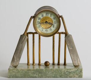Cricket clock. A Victorian brass mantel clock. An attractive late Victorian/ Edwardian brass clock of cricketing interest, formed with a central drum form enamelled dial and workings attached two sets of cricket stumps and bails with cricket bats leaning 