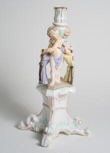 Cricket themed candlestick. Large ceramic candlestick the centre features two winged cherubs holding cricket bats on plinth, the base with four feet. A very colourful design in pastel coloured glaze in pink, light blue, mauve and yellow with gilt lustre.