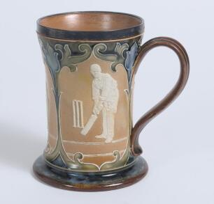 Doulton Lambeth stoneware waisted ceramic tankard with three moulded relief vignettes of cricketers, a batsman, bowler and wicket keeper in white on brown background. With stylised floral leaf and flower decoration to top and beneath in green and light bl
