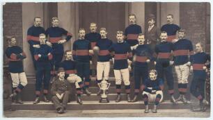 Rugby Union. Wakefield Trinity Rugby Football Club c.1879. Large and early original mono photograph heightened in club colours of red and blue of the Wakefield Trinity team with the Yorkshire Challenge Cup trophy, standing on the stone steps of what appea