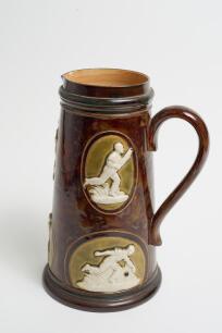 Cricketing jug. A large and impressive Doulton Lambeth stoneware tapering jug in a dark brown mottled colour, moulded in relief with six raised figures, three of batsman, two of fielders and a wicket keeper in roundels, all different and in various positi