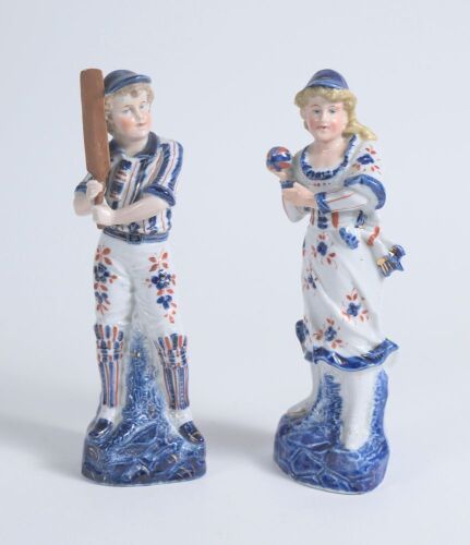 Cricket figures. A pair of German porcelain figures, the boy holding a cricket bat, wearing pads with a cap, the girl in three quarter length dress wearing a cap, she is holding a ball. Each figure is beautifully decorated in blue, red and gold lustre. 9&rdquo;