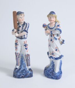 Cricket figures. A pair of German porcelain figures, the boy holding a cricket bat, wearing pads with a cap, the girl in three quarter length dress wearing a cap, she is holding a ball. Each figure is beautifully decorated in blue, red and gold lustre. 9&rdquo;