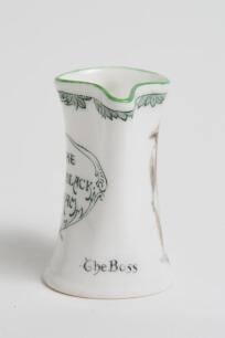 &lsquo;The Boss&rsquo;. Royal Doulton Black Boy miniature flared lip jug, with printed image of a boy in red shirt and a floppy hat, wearing an umpire&rsquo;s coat with bat to side, title to side. To verso a crest with &lsquo;The All Black Team&rsquo;. Green floral decoration to outer